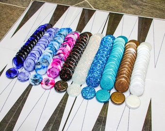 May include: A backgammon set with a variety of colorful game pieces. The pieces are arranged in rows, showcasing a spectrum of colors including blue, pink, black, white, and brown. The game board has a white surface with black triangular markings.