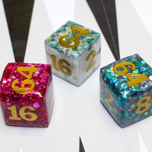 May include: Three glittery dice with gold numbers. The dice are pink, blue, and white with gold glitter embedded in the resin. The numbers on the dice are 4, 6, 16, 2, and 8.