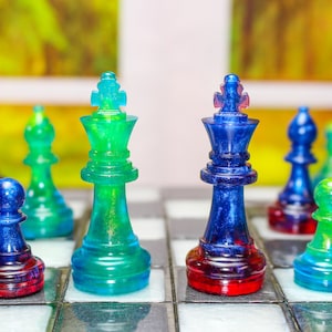 May include: A close-up of a chessboard with two kings facing each other. The king on the left is green and the king on the right is blue. The chess pieces are made of glass and have a translucent appearance.