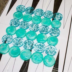 May include: Backgammon game pieces in teal and white. The teal pieces are solid, while the white pieces have a teal speckled design. The pieces are arranged on a backgammon board with black and white triangles.