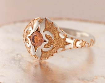 May include: A silver ring with a gold-toned design featuring a star and crescent moon motif. The ring has a large, round centre stone with a brown hue.