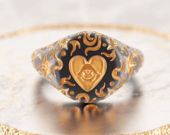 May include: A black signet ring with a gold heart design featuring an eye inside. The ring is decorated with a swirling pattern of stars and other abstract shapes.