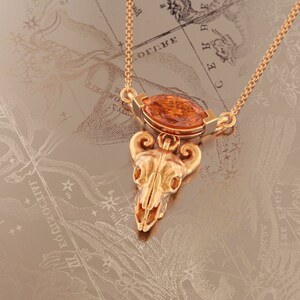 May include: Gold necklace with a pendant featuring a detailed skull with horns and an orange gemstone. The necklace is on a background of a vintage map.