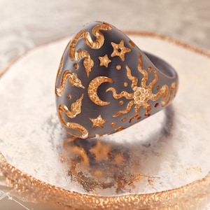 May include: A black ring with a gold design of a sun, moon, and stars. The ring is on a wooden surface.