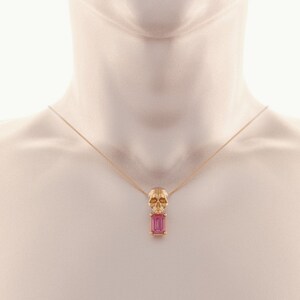 Puede incluir: Un collar dorado con un colgante de calavera y una gema rectangular rosa. La calavera tiene detalles faciales. El collar se muestra en un maniqu&iacute;.