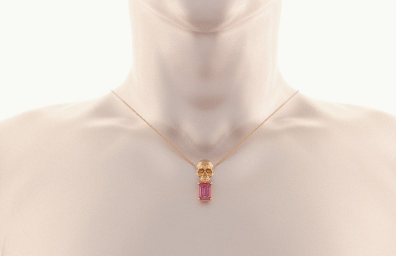 Puede incluir: Un collar dorado con un colgante de calavera y una gema rectangular rosa. La calavera tiene detalles faciales. El collar se muestra en un maniqu&iacute;.