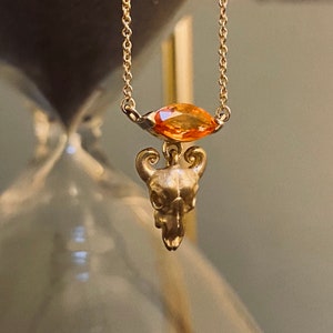 May include: A gold chain necklace with a small, detailed skull pendant and a large, orange gemstone.