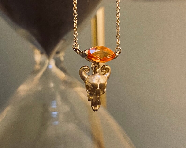 May include: A gold chain necklace with a small, detailed skull pendant and a large, orange gemstone.