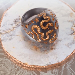 May include: A black ring with a gold snake design. The ring is set on a white and gold surface.