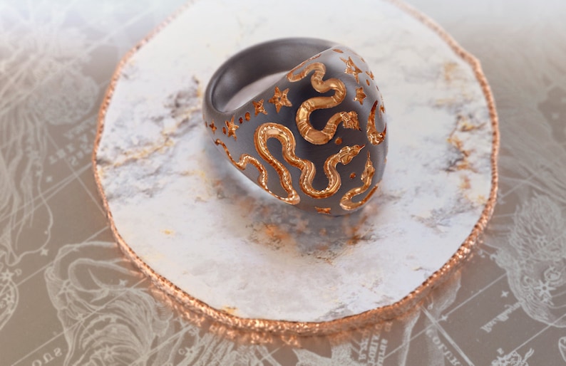 May include: A black ring with a gold snake design. The ring is set on a white and gold surface.