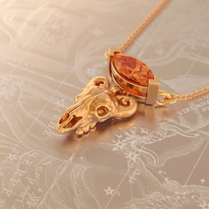 May include: A gold necklace with a pendant featuring a detailed ram skull and a large, orange gemstone. The necklace is on a background of a star chart.