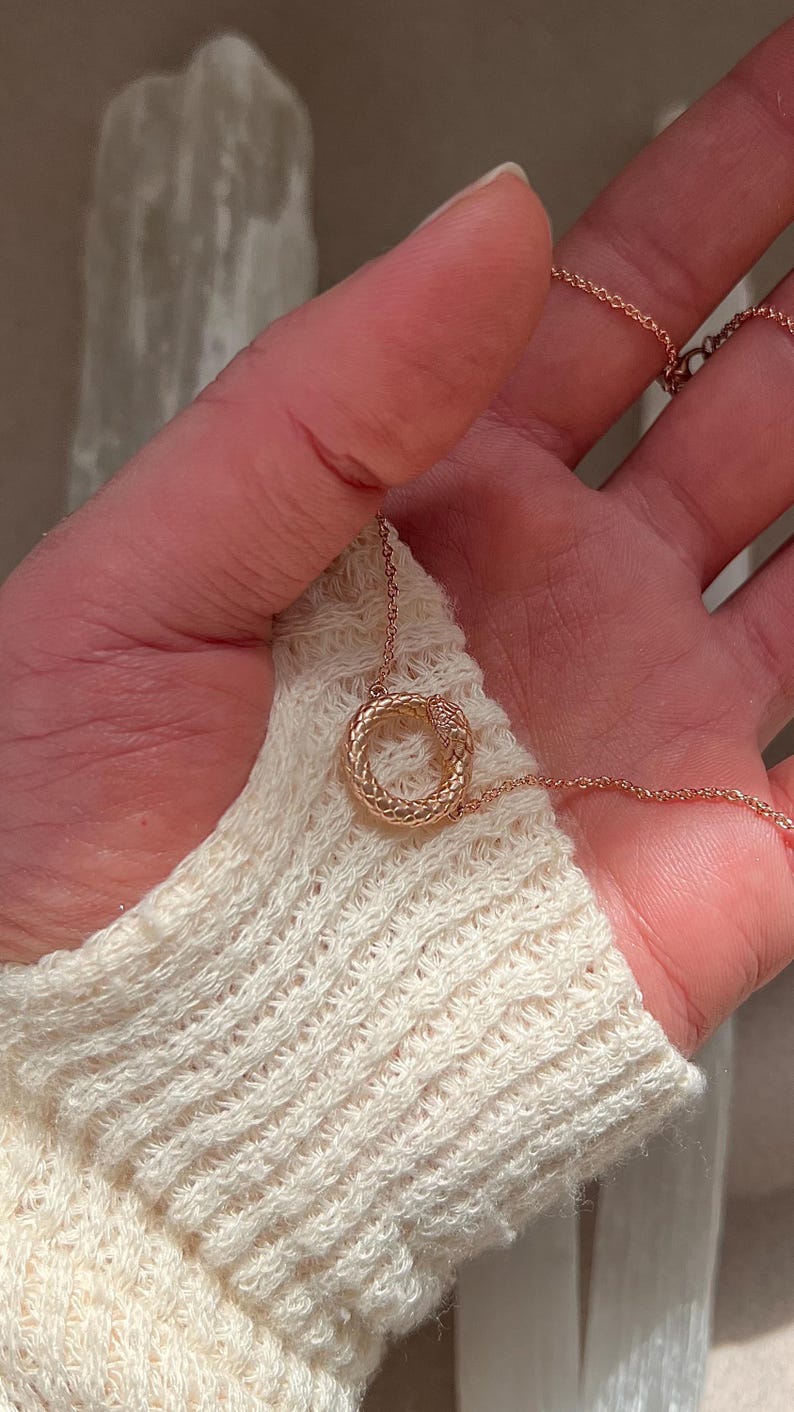 May include: A rose gold necklace featuring a snake-shaped pendant. The pendant is a circle with a snake wrapped around it.