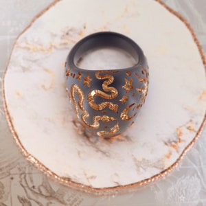 May include: A black ring with a gold snake and star design. The ring is on a white marble surface.