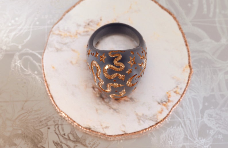 May include: A black ring with a gold snake and star design. The ring is on a white marble surface.