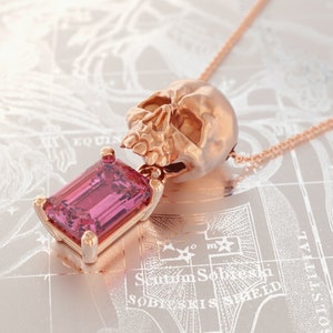 Puede incluir: Collar con colgante de calavera en oro rosa con una gran gema rosa rectangular. La calavera es detallada y la gema est&aacute; engastada en un marco dorado. El collar se muestra sobre un fondo similar a un mapa.