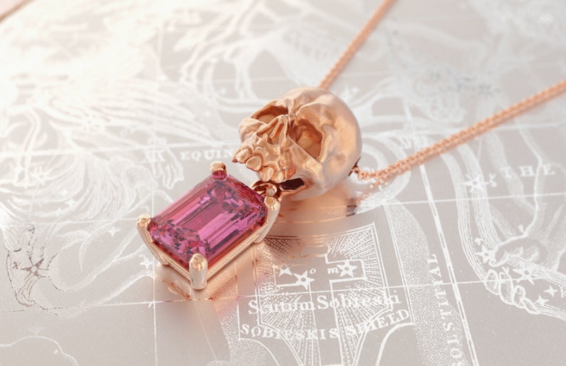 Puede incluir: Collar con colgante de calavera en oro rosa con una gran gema rosa rectangular. La calavera es detallada y la gema est&aacute; engastada en un marco dorado. El collar se muestra sobre un fondo similar a un mapa.