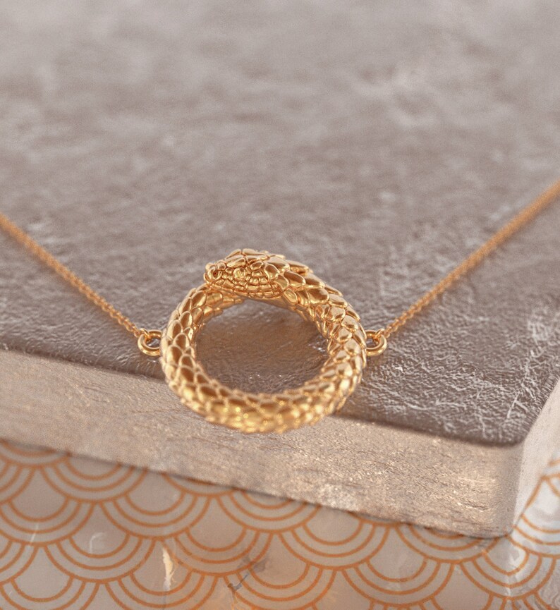 May include: A gold snake necklace with a snake head forming a circle. The snake is detailed with scales and has a smooth, shiny finish.
