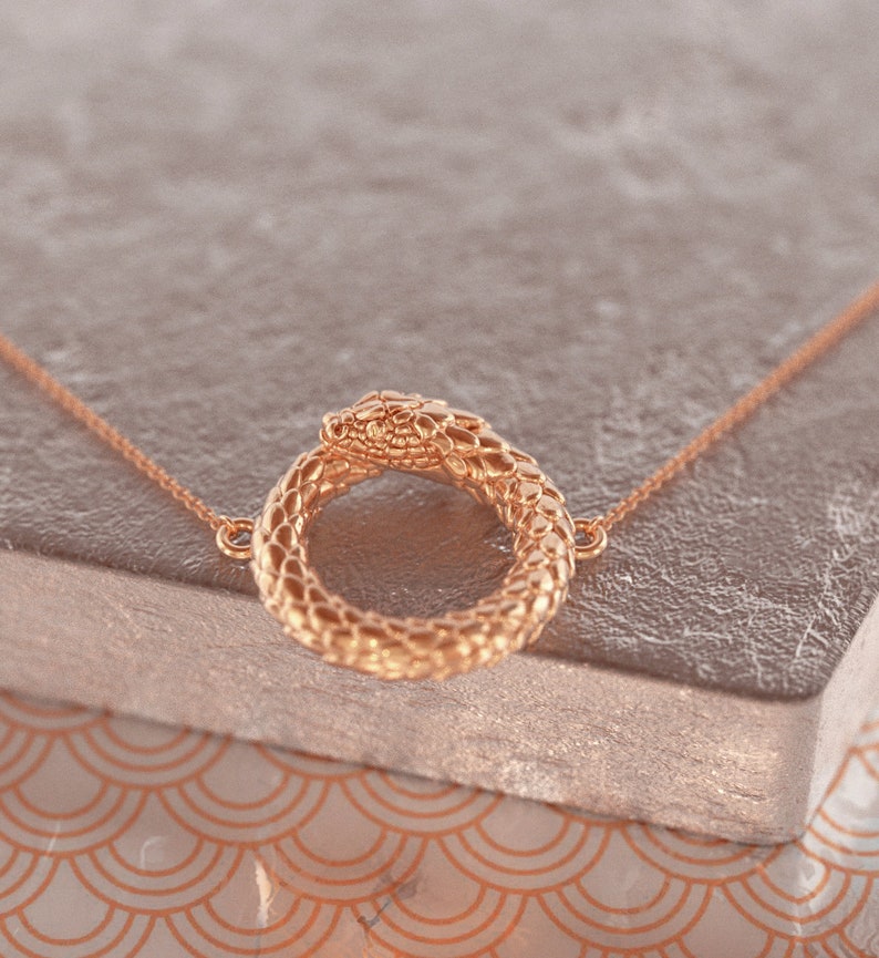May include: A gold snake necklace with a circular pendant. The snake is coiled into a circle with its head facing the viewer.