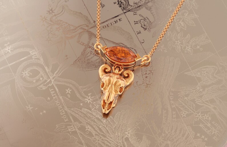 May include: Gold necklace with a pendant featuring a detailed skull with horns and an orange gemstone. The necklace is on a background of a vintage map.
