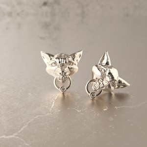 Puede incluir: Dos pendientes de botón plateados con forma de cabeza de zorro. Cada pendiente presenta una cabeza de zorro detallada con un anillo en la boca, que sostiene una pequeña piedra redonda y transparente. Sobre una superficie gris claro.