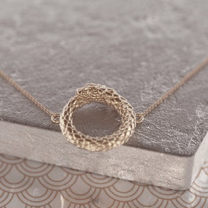 May include: A gold snake necklace with a snake head forming a circle. The necklace is on a grey textured surface.