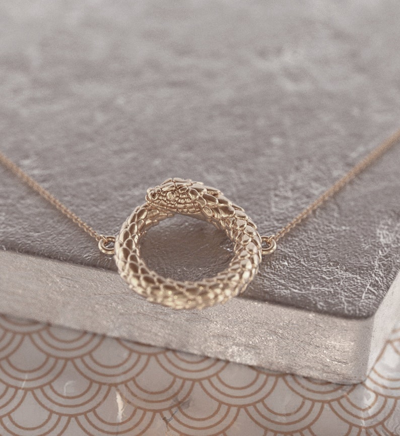 May include: A gold snake necklace with a snake head forming a circle. The necklace is on a grey textured surface.