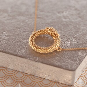 May include: A gold snake necklace with a circular pendant. The snake is curled into a circle, with its head at the top of the circle. The necklace is on a light brown surface.