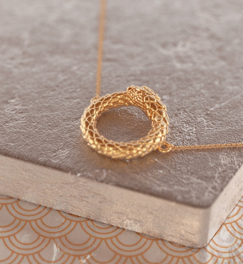 May include: A gold snake necklace with a circular pendant. The snake is curled into a circle, with its head at the top of the circle. The necklace is on a light brown surface.