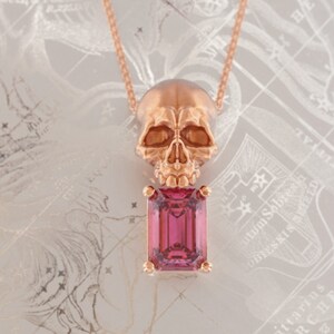 Puede incluir: Un collar con colgante de oro rosa con un colgante de calavera sobre una gema rosa rectangular. El collar cuelga de una delicada cadena. El fondo tiene un dise&ntilde;o claro y estampado.