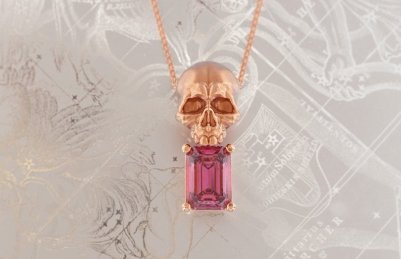 Puede incluir: Un collar con colgante de oro rosa con un colgante de calavera sobre una gema rosa rectangular. El collar cuelga de una delicada cadena. El fondo tiene un dise&ntilde;o claro y estampado.