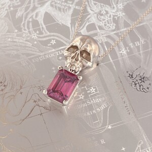 Puede incluir: Un collar con colgante de plata con un dise&ntilde;o de calavera sobre una gema rosa rectangular. El colgante cuelga de una delicada cadena. El fondo es una superficie gris claro con un dise&ntilde;o de mapa celestial.