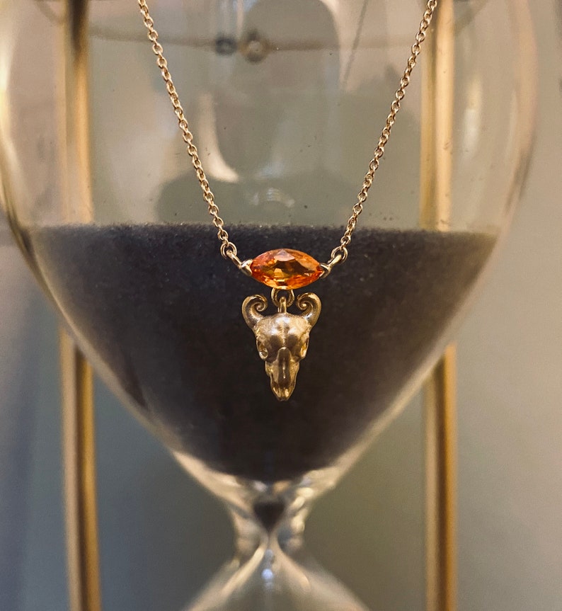May include: A gold chain necklace with a small, detailed skull pendant and a large, orange gemstone. The necklace is hanging from a glass hourglass.