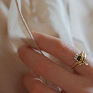 May include: A gold ring with a black oval gemstone surrounded by smaller white gemstones. The ring is set on a finger wearing a second gold band.