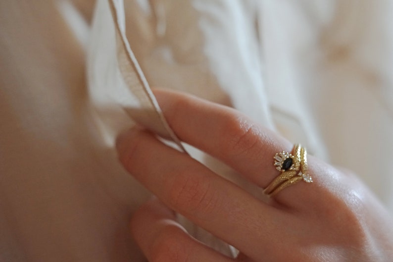 May include: A gold ring with a black oval gemstone surrounded by smaller white gemstones. The ring is set on a finger wearing a second gold band.