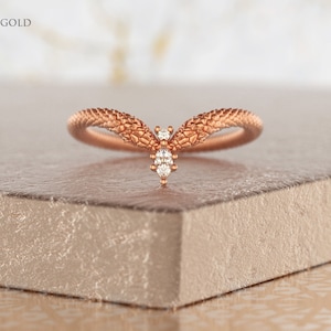 May include: A rose gold ring with a textured band and a small diamond accent. The ring is shaped like a V and is set on a light brown surface.