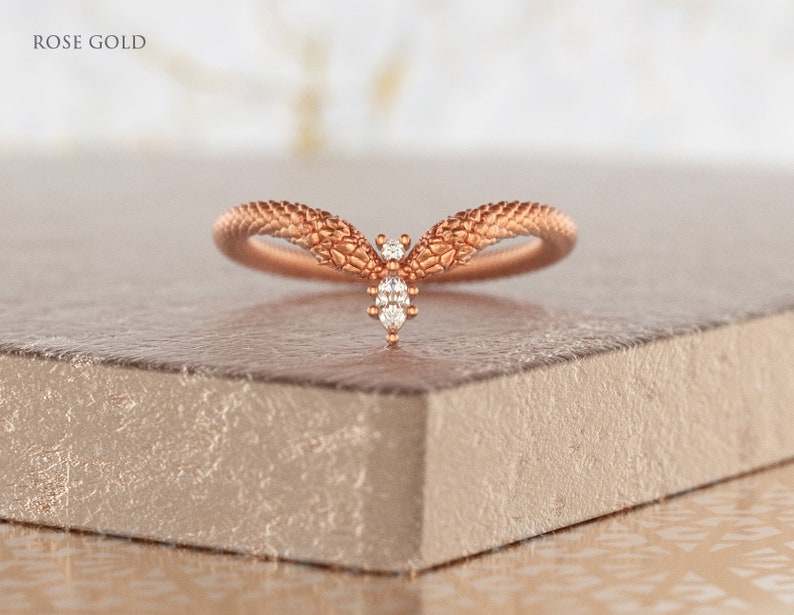 May include: A rose gold ring with a textured band and a small diamond accent. The ring is shaped like a V and is set on a light brown surface.