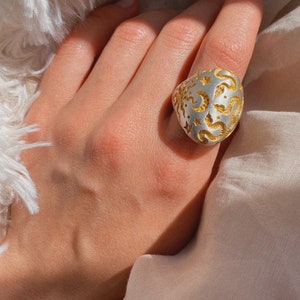 May include: A silver ring with a large, oval-shaped face. The face of the ring is decorated with a detailed pattern of gold stars, moons, and swirls.