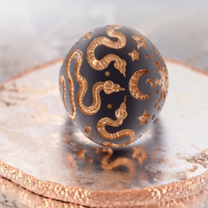 May include: A black sphere with a gold snake and star pattern. The sphere is sitting on a silver and gold circular stand.