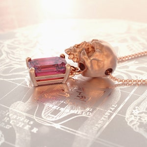 Puede incluir: Collar con colgante de oro rosa con una calavera y una gema rosa rectangular. La calavera sostiene la gema. La cadena del collar tambi&eacute;n es de oro rosa.