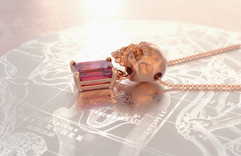 Puede incluir: Collar con colgante de oro rosa con una calavera y una gema rosa rectangular. La calavera sostiene la gema. La cadena del collar tambi&eacute;n es de oro rosa.
