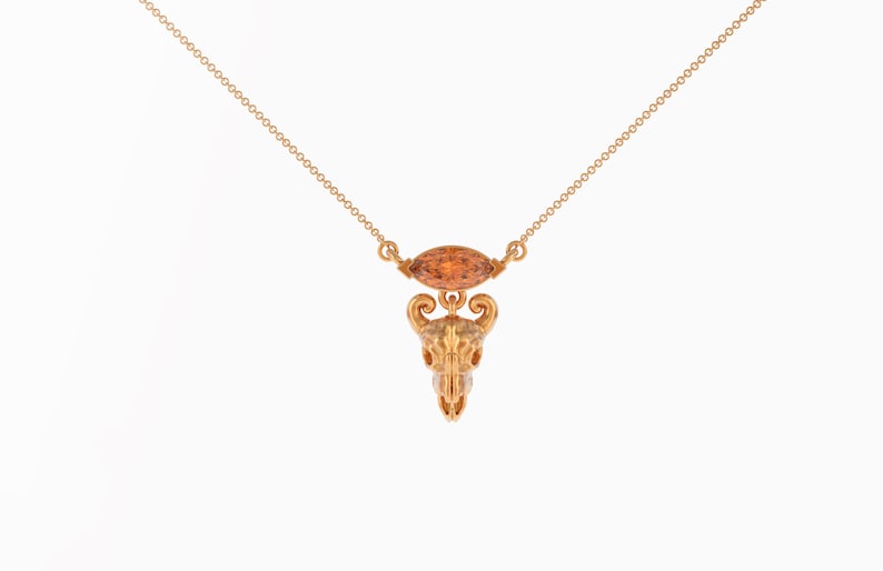 May include: A gold necklace with a detailed skull pendant and a small, orange gemstone.