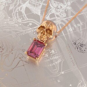 Puede incluir: Collar con colgante de calavera dorada con una gema rosa rectangular. La calavera es detallada y la gema est&aacute; engastada en un marco dorado. El collar descansa sobre una superficie gris claro con un dise&ntilde;o de mapa estelar.