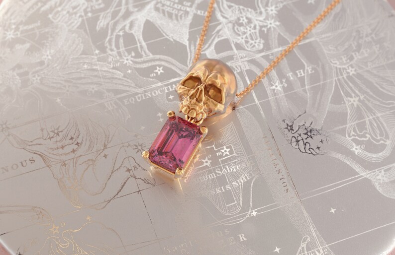 Puede incluir: Collar con colgante de calavera dorada con una gema rosa rectangular. La calavera es detallada y la gema est&aacute; engastada en un marco dorado. El collar descansa sobre una superficie gris claro con un dise&ntilde;o de mapa estelar.