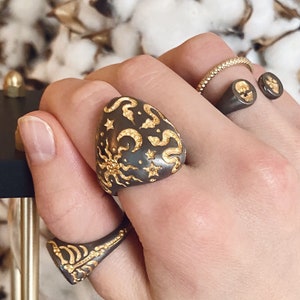 May include: A hand wearing three rings. The ring on the index finger is a large, dark grey ring with a gold design of stars, moons, and swirls. The ring on the middle finger is a thin gold band. The ring on the little finger is a dark grey ring with two gold skulls.