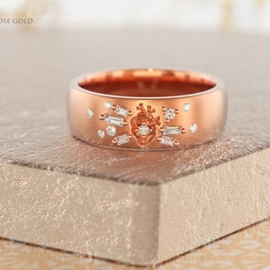 May include: A rose gold band ring with a heart design and small diamonds. The ring is on a textured surface.