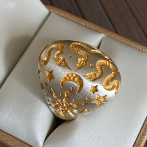 May include: A silver ring with a gold design of snakes, stars, and crescent moons. The ring is in a white box.