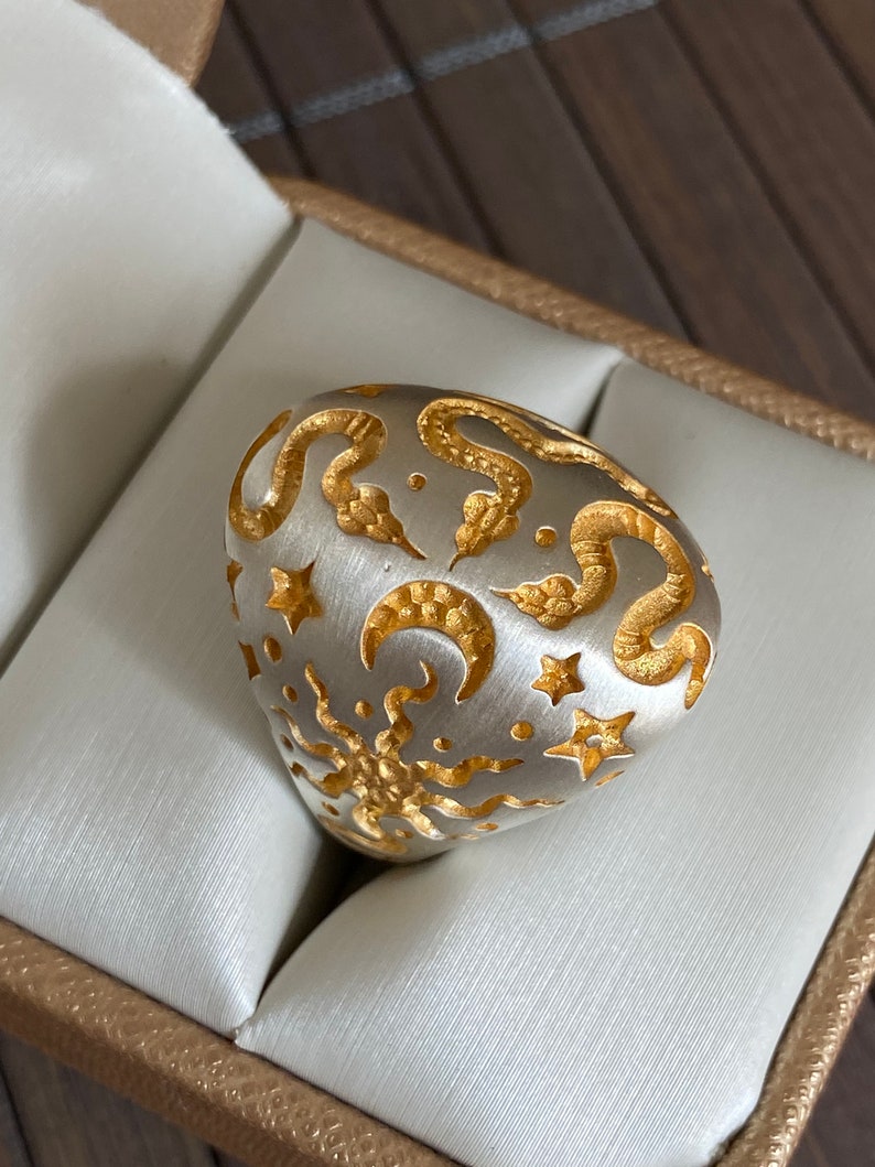 May include: A silver ring with a gold design of snakes, stars, and crescent moons. The ring is in a white box.