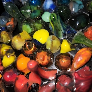 Op de afbeelding: Een verzameling kleurrijke glazen knikkers in verschillende maten en vormen. De knikkers tonen een reeks kleuren, waaronder blauw, groen, geel, oranje en rood. Sommige knikkers hebben wervelende patronen, andere zijn effen.