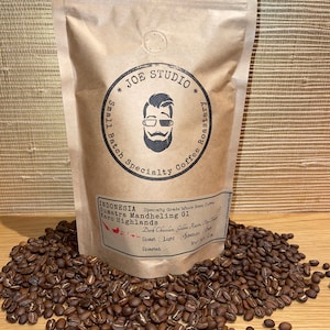 May include: A brown paper bag of whole bean coffee from Joe Studio, Sumatra Mandheling G1, Karo Highlands. The bag is surrounded by roasted coffee beans. The bag has a circular logo with a bearded man wearing glasses.