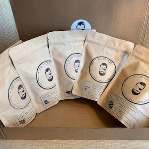 May include: Six brown paper bags with different coffee blends. Each bag has a logo with a man's face and the text "Joe Studio Small Batch Specialty Coffee Roasters". The bags are labeled with the origin of the coffee beans, including Ethiopia, Colombia, Honduras, Tanzania, and Kenya.